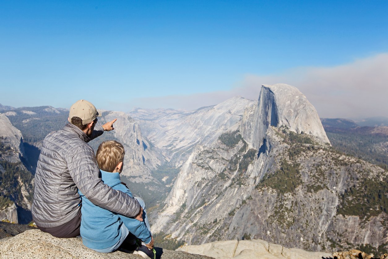 Back view of family of two, father and son, sitting together at glacier point and enjoying view of half dome and yosemite valley in yosemite national park, california, family adventure vacation concept